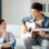 A young Asian man plays sits on a couch in a living room and plays an acoustic guitar to a young asian woman listening happily.