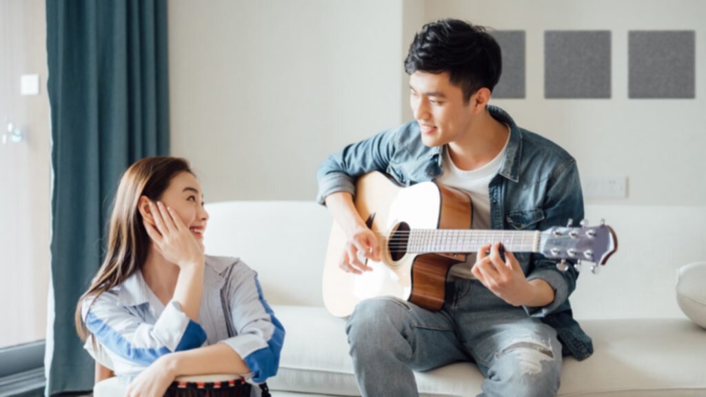 A young Asian man plays sits on a couch in a living room and plays an acoustic guitar to a young asian woman listening happily.